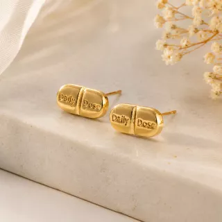 14K gold plated Daily Dosage Pill Stud Earrings displayed on a minimalist marble surface, showcasing the polished capsule design engraved with "Daily Dose".