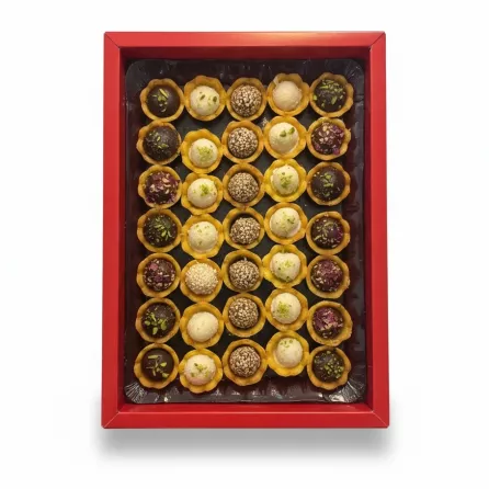 Assorted small round sweets in yellow cups in a red box, topped with sesame seeds, pistachio, and rose petal decorations