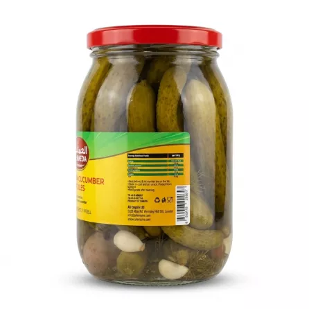 A clear glass jar filled with whole Al Maeda Premium Cucumber Pickles, visible dill sprigs, and garlic cloves, presented from a side angle. A large yellow label displays a detailed nutritional values table, company information, and a barcode, all on a clean, isolated background.