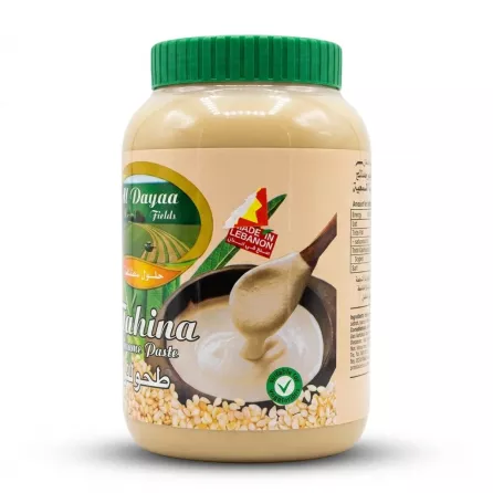 A 1:1 e-commerce photo of a clear plastic jar of Al Dayaa Tahina Sesame Paste, featuring its front label with a "Made in Lebanon" sticker, a wooden spoon scooping creamy tahina, and scattered sesame seeds. The product is perfectly clean, in sharp focus, and centered against a plain white background with professional, even studio lighting and a soft, symmetrical contact shadow.