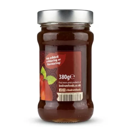 A clear glass jar filled with Bodrum Rosehip Jam is centered, showing the back label. The label states 'no added colouring or flavouring', '380g e', the website 'bodrumfoods.co.uk', social media handles, and a barcode. The product is against a plain white background with professional studio lighting and a soft, even shadow.