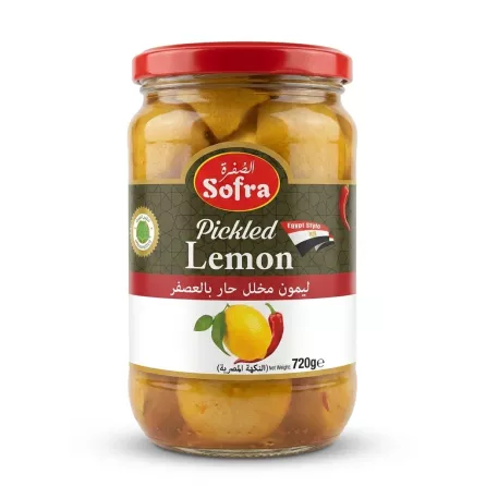 A 720g glass jar of Sofra Pickled Lemon Egyptian Style with a red lid, featuring a green label with English and Arabic text and an illustration of lemons and a red chili. The jar is centered on a clean white background. PRODUCT TITLE: Sofra Pickled Lemon Egyptian Style PRODUCT: A professional, studio-quality e-commerce photograph of a 720g glass jar of Sofra Pickled Lemon Egyptian Style, as seen in image_0.png. The jar is centered and fills approximately 85% of the frame. All background elements (hand, store shelves, etc.) have been completely removed. The jar stands on a pristine, seamless white surface. The product label is meticulously cleaned, retaining all factual information: the 'Sofra' logo, 'Pickled Lemon' text, the red 'Egypt Style' band, the full Arabic text "ليمون مخلل حار بالعصفر" and "النكهة المصرية", the image of the lemons and red chili, and the '720g e' weight. Crucially, the yellow and red price sticker ('£1.69' and arrow) and the small green 'Hot' sticker have been seamlessly removed, revealing the clean, continuous label paper and text behind them. The jar is lit with soft, diffused professional studio lighting that is even across the entire product, with no harsh highlights or uneven shadows. Soft, equal-intensity shadows are cast on both the left and right sides of the base of the jar, giving it a stable presence. The glass texture is rendered clearly, showing the liquid brine and whole pickled lemons inside. The focus is tack-sharp on the central label and the front of the jar. The aspect ratio is 1:1 square.