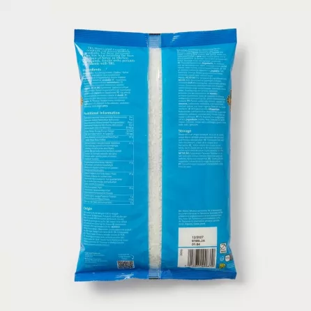 A professional studio photograph showcasing the back of the TRS Desiccated Coconut Medium pack. The bright blue packaging is clean, well-lit, and centered on a seamless light grey background, prominently displaying the multi-lingual ingredients, detailed nutritional information table, and specific batch details including the 12/2027 best before date and barcode. The clear central window reveals the shredded coconut inside.