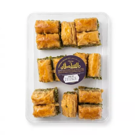 A professional, centered studio photograph of a clear plastic tray containing a selection of assorted Sultan baklava pieces, featuring layers of filo and nut fillings. The central circular label is clearly visible with Arabic text and a use-by date of 28/2026, set against a seamless white background.