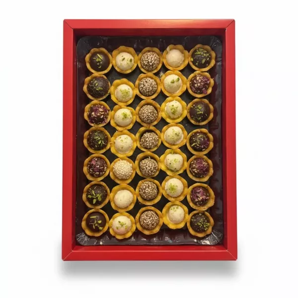 Assorted small round sweets in yellow cups in a red box, topped with sesame seeds, pistachio, and rose petal decorations