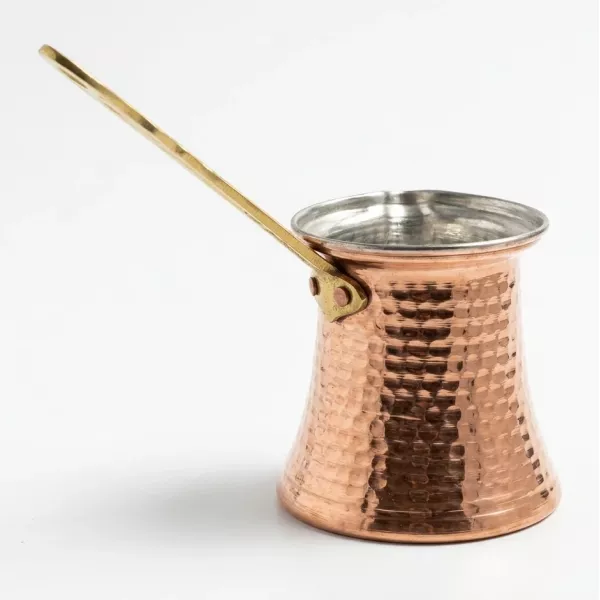 A close-up studio photograph of a traditional handcrafted copper Turkish coffee pot, featuring a distinctive hammered texture and a long, polished brass handle, set against a seamless white background.