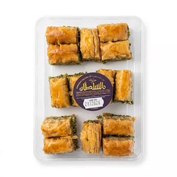 A professional, centered studio photograph of a clear plastic tray containing a selection of assorted Sultan baklava pieces, featuring layers of filo and nut fillings. The central circular label is clearly visible with Arabic text and a use-by date of 28/2026, set against a seamless white background.