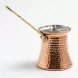 A close-up studio photograph of a traditional handcrafted copper Turkish coffee pot, featuring a distinctive hammered texture and a long, polished brass handle, set against a seamless white background.